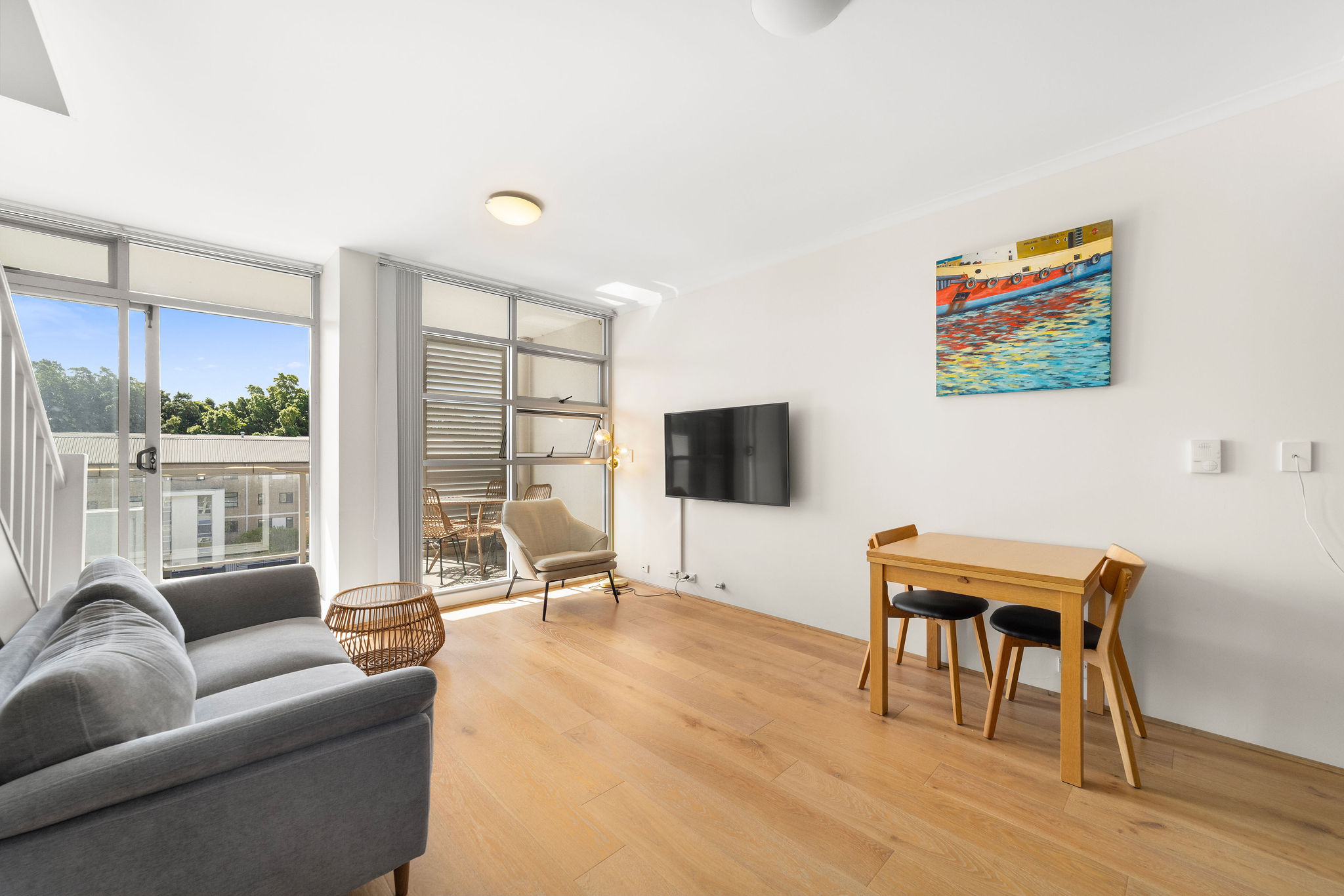 Stylish furnished loft living area with grey sofa, wooden dining table, and balcony access, featuring natural light and views of tranquil gardens.